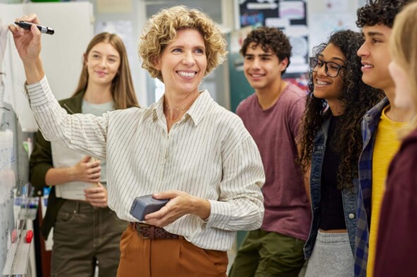 a female teacher writing on a whiteboard while smiling at a diverse group of five teens gathered around her