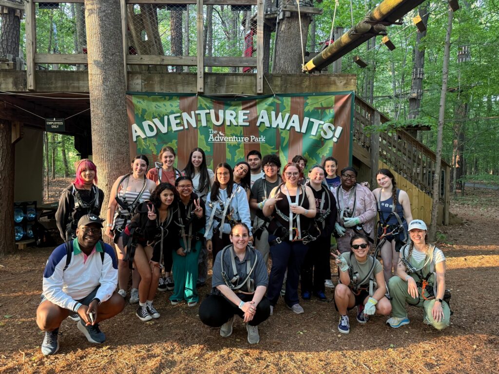 group of older teens and adults posing for a photo in a wooded setting in front of a sign that reads "Adventure Awaits"