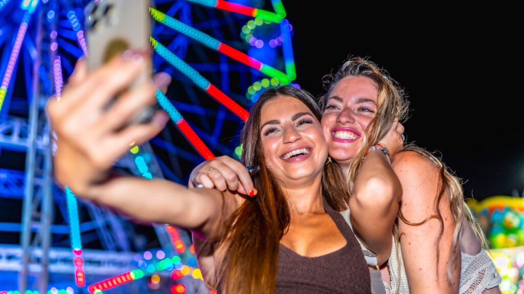 2 older teen girls smiling for a selfie in front of a colorful Ferris wheel at a senior grad night