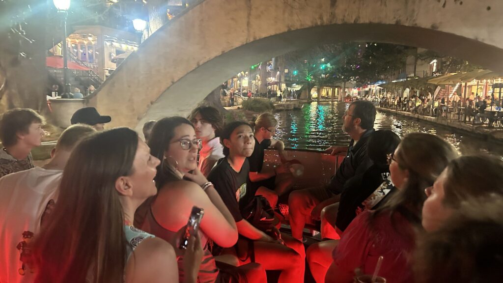 a group of students in an open boat floating under an arched bridge on the San Antonio River Walk
